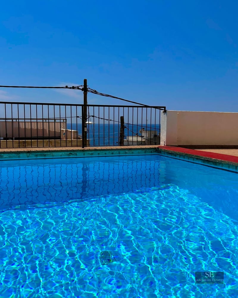 Clear blue rooftop swimming pool with a black railing overlooking the sea under a bright blue sky.