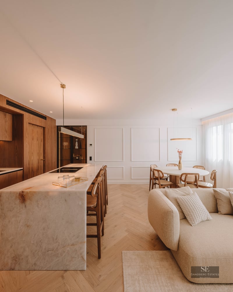 Modern open-plan room with a marble kitchen island, wood cabinets, herringbone floors, and a large cream sectional sofa.