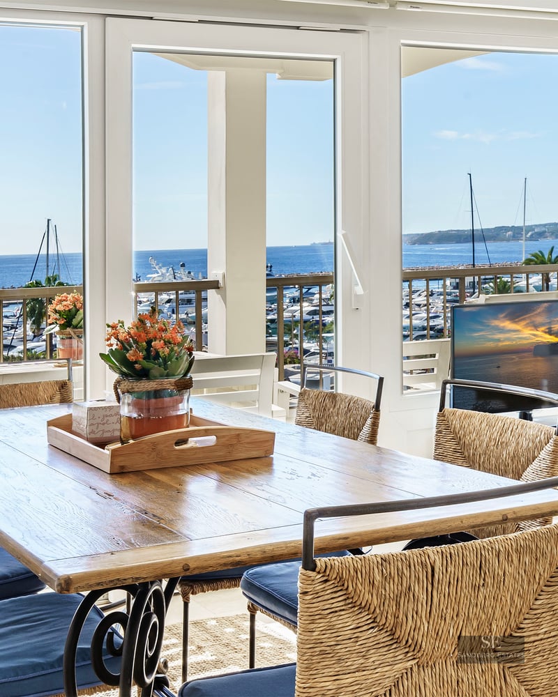 Sunlit dining room with a wooden table and woven chairs looking out over a yacht-filled marina and the blue ocean.