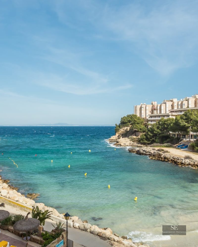 High-angle view of a turquoise sea cove with a small beach, stone walkways, and coastal apartment buildings.