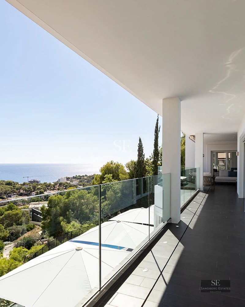 A long white modern balcony with a glass railing overlooking green trees and the blue sea under a clear sky.