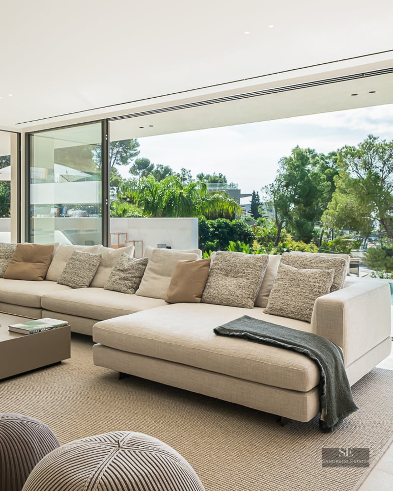 A bright, modern living room with a large cream sectional sofa looking out through glass walls to a private pool and trees.
