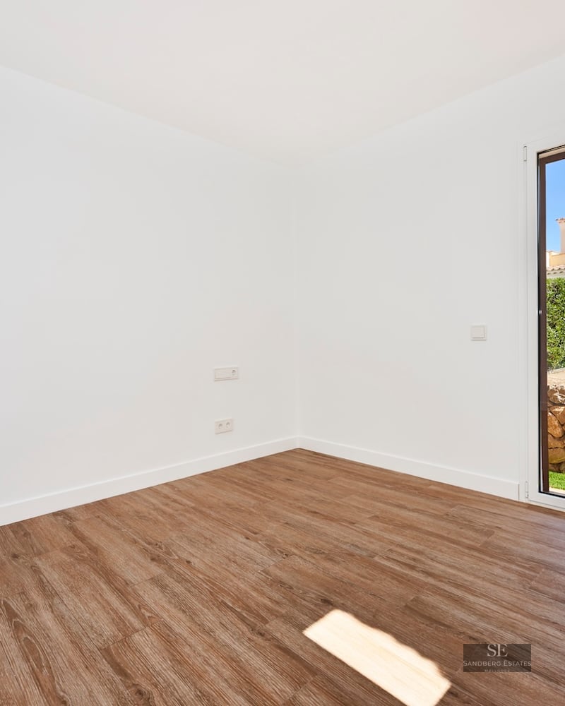 Empty room with white walls, wood floors, and a glass door opening to a garden with a stone wall.