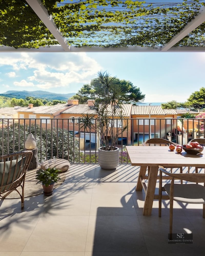 Sunlit terrace with wooden dining table, rattan chair, and vine-covered pergola overlooking a Mediterranean landscape.