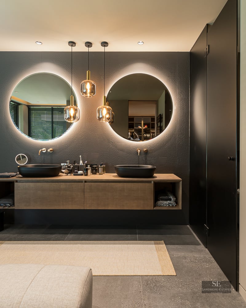 Modern master bathroom featuring two round backlit mirrors, black vessel sinks on a wooden vanity, and grey stone floors.