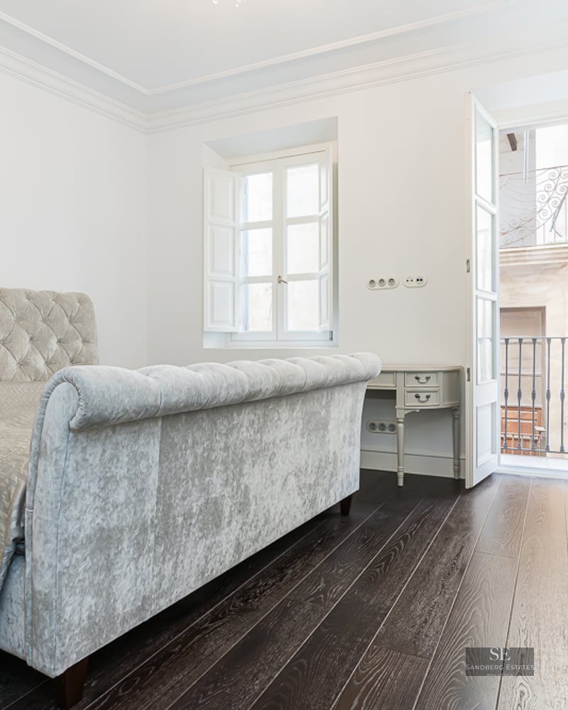 Bright bedroom featuring a grey velvet tufted bed, dark wood floors, and a balcony with wrought iron railing.