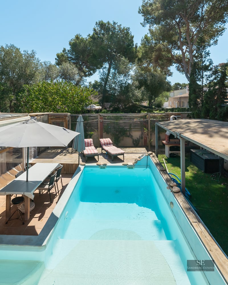 Rectangular turquoise pool surrounded by wooden decking, outdoor dining area, and lush trees under a clear blue sky.