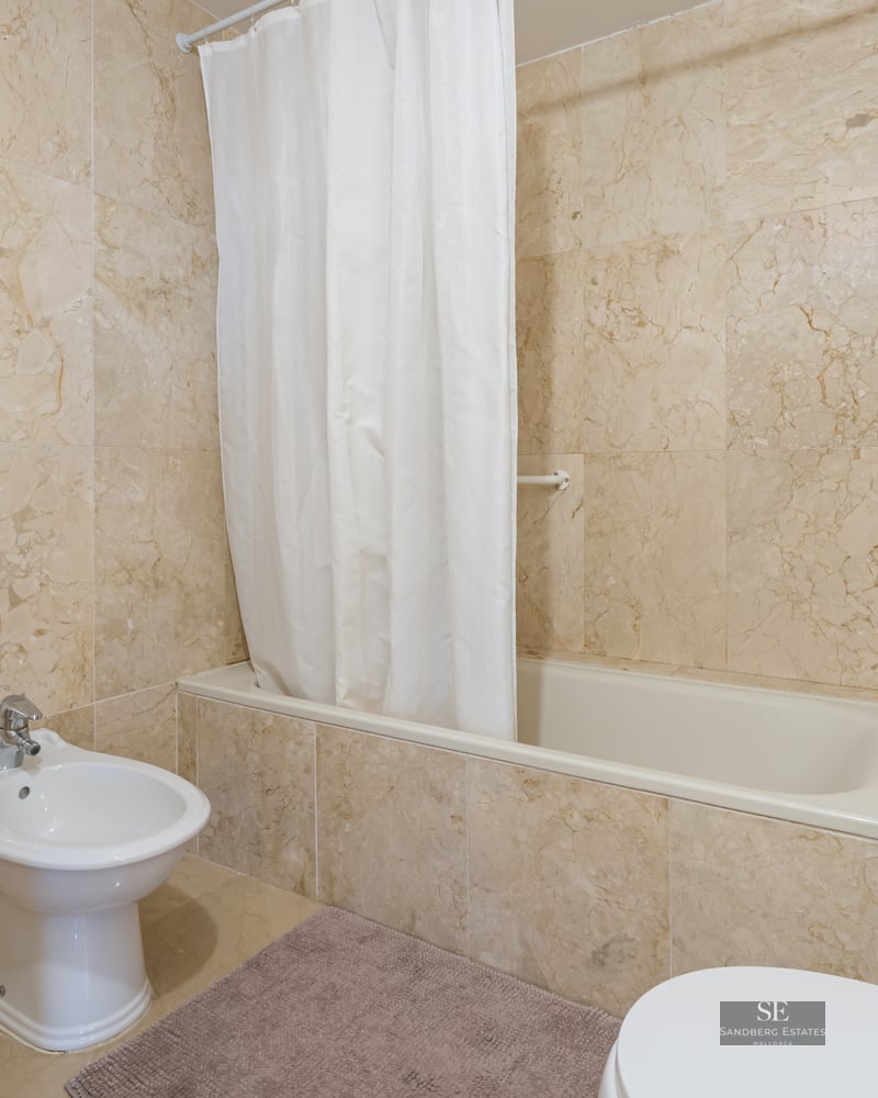 Bathroom featuring beige marble walls, a white bathtub with curtain, and a white ceramic bidet.