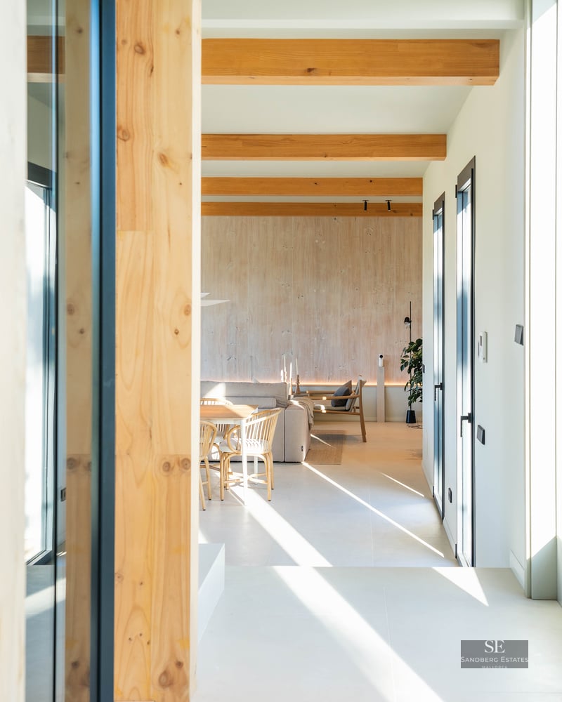 Modern living room featuring exposed wooden beams, beige sofa, and strong sunlight through large windows.