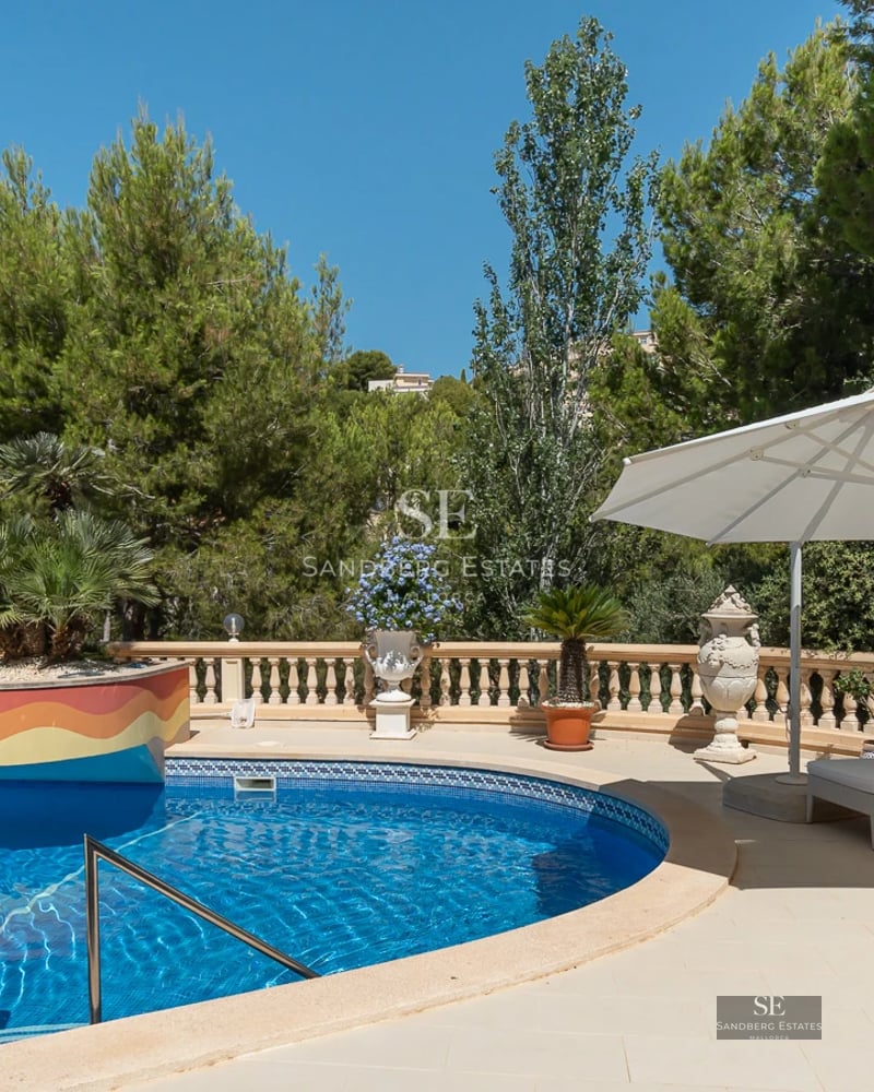 Image of a modern villa facade with reflective pool, surrounded by greenery and a bright blue sky.