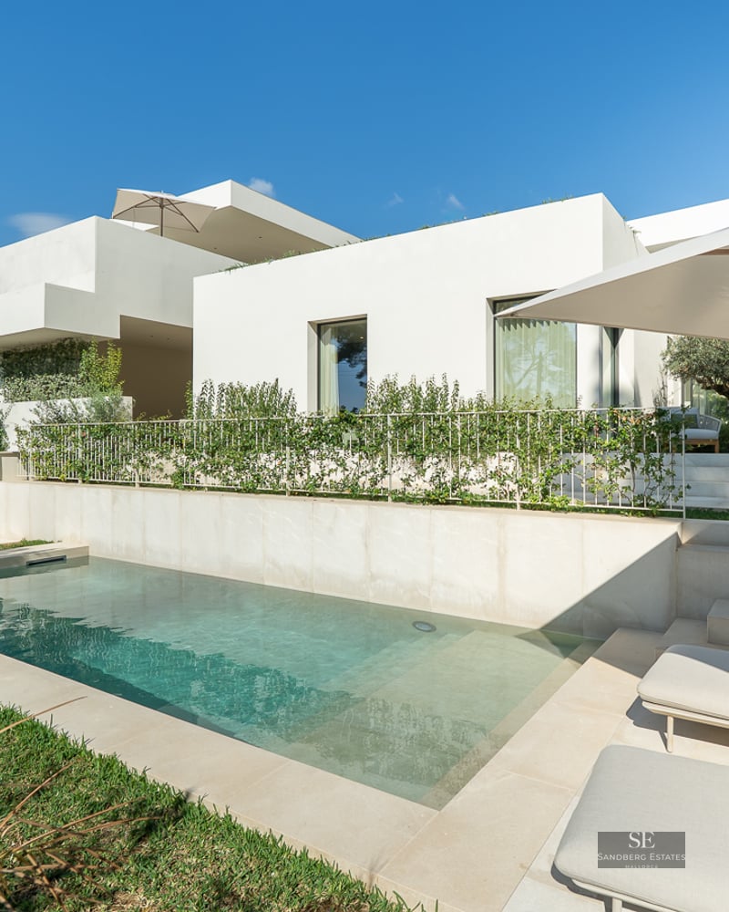 Modern white villa with a rectangular swimming pool, stone patio, sun loungers, and lush green landscaping under a blue sky.