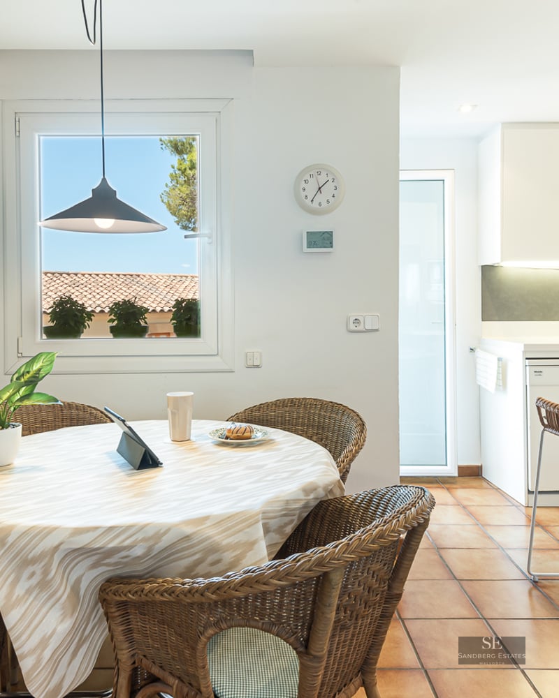Round dining table with wicker chairs and terracotta flooring next to a modern white kitchen.