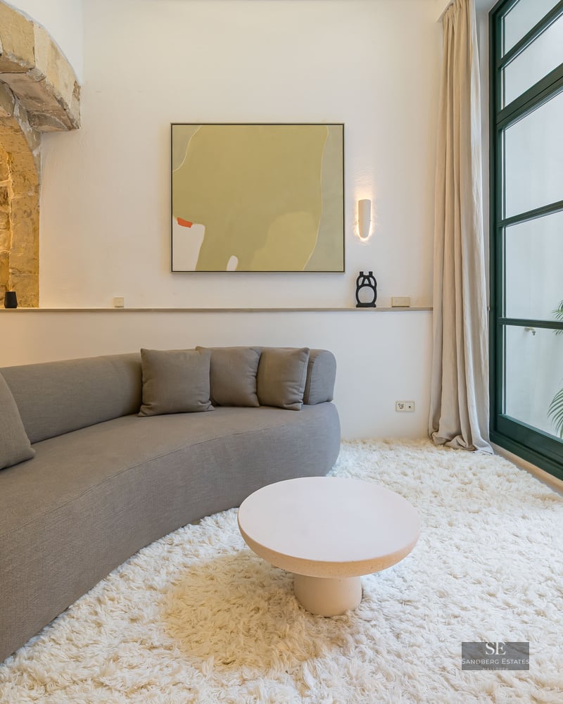 A modern living room featuring a curved taupe sofa, shaggy white rug, stone archway, and floor-to-ceiling glass doors to a patio.