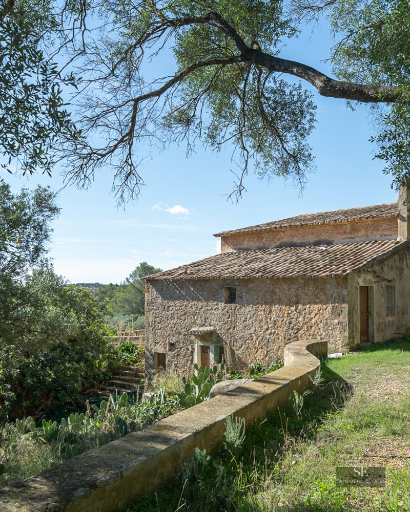 A historic two-story stone house with a terracotta roof nestled among trees under a clear blue sky.