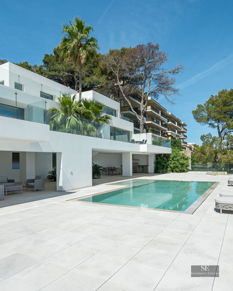Modern white villa with a rectangular swimming pool, grey sun loungers, and glass railings under a clear blue sky.