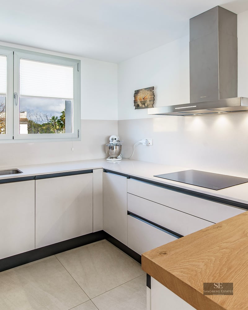 L-shaped modern white kitchen with wood breakfast bar, stainless steel appliances, and a large window.