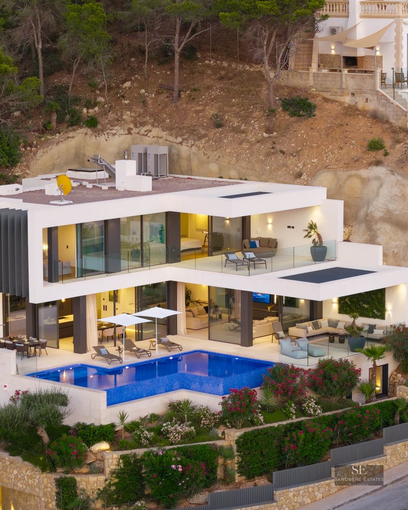 Modern white villa on a hillside featuring a bright blue swimming pool and glass-walled terraces.