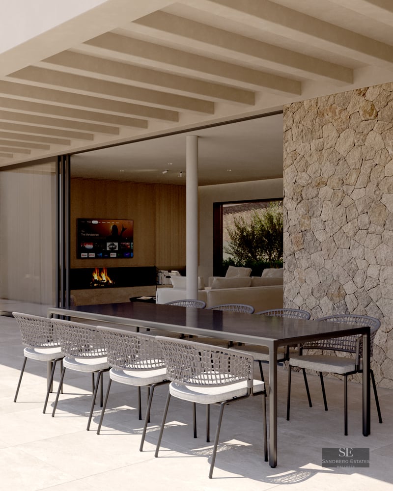 A modern covered terrace with a large black dining table, woven chairs, natural stone wall, and sliding glass doors.