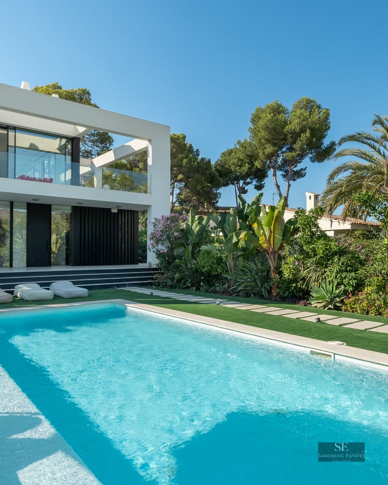 Modern white two-story villa with a large rectangular swimming pool, lush green garden, and outdoor dining area.