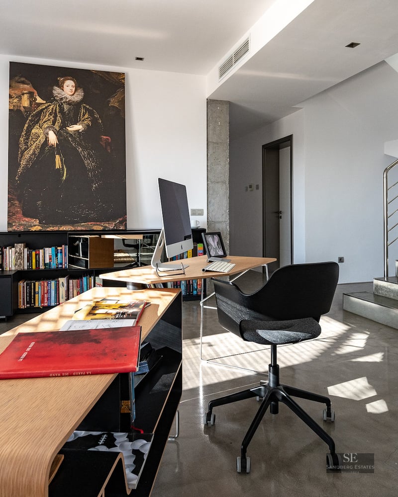 Modern home office with a curved wooden desk, black ergonomic chair, large classical painting, and concrete stairs.