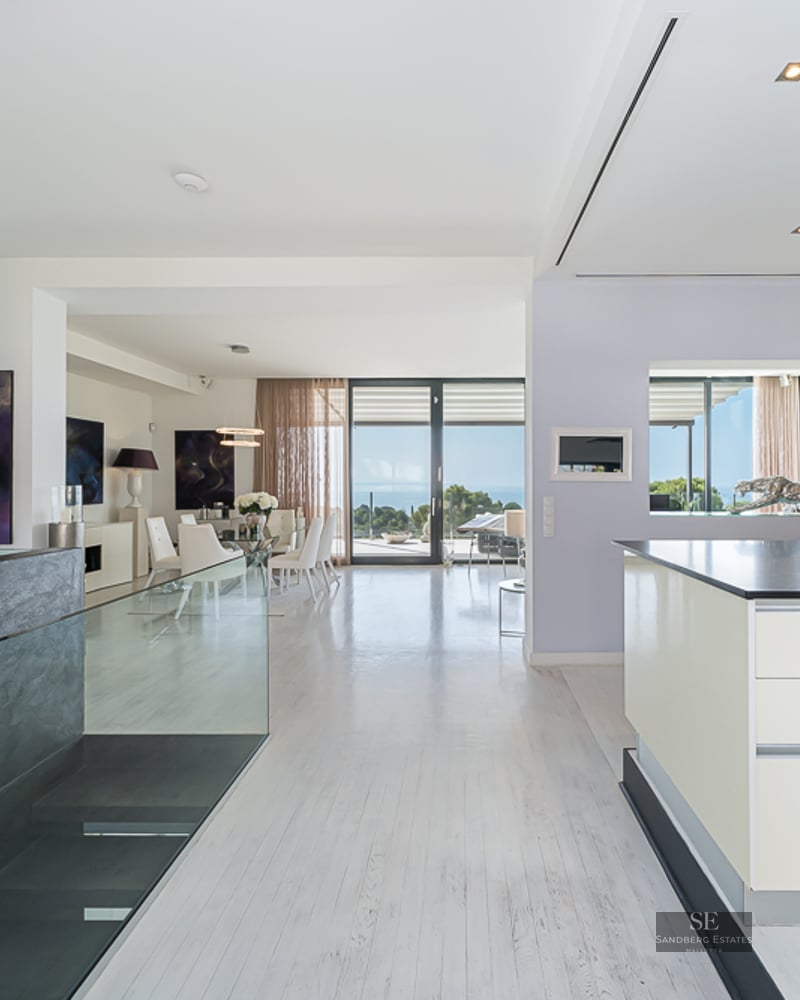 Modern open-plan interior featuring a white kitchen island, glass dining table, and large windows with sea views.