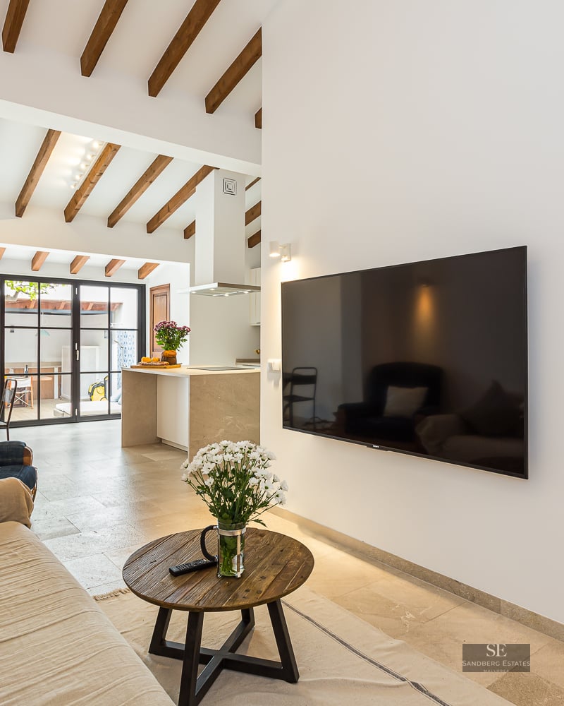 Modern living room featuring a beige sofa, wooden ceiling beams, large flat-screen TV, and a view into an open kitchen.