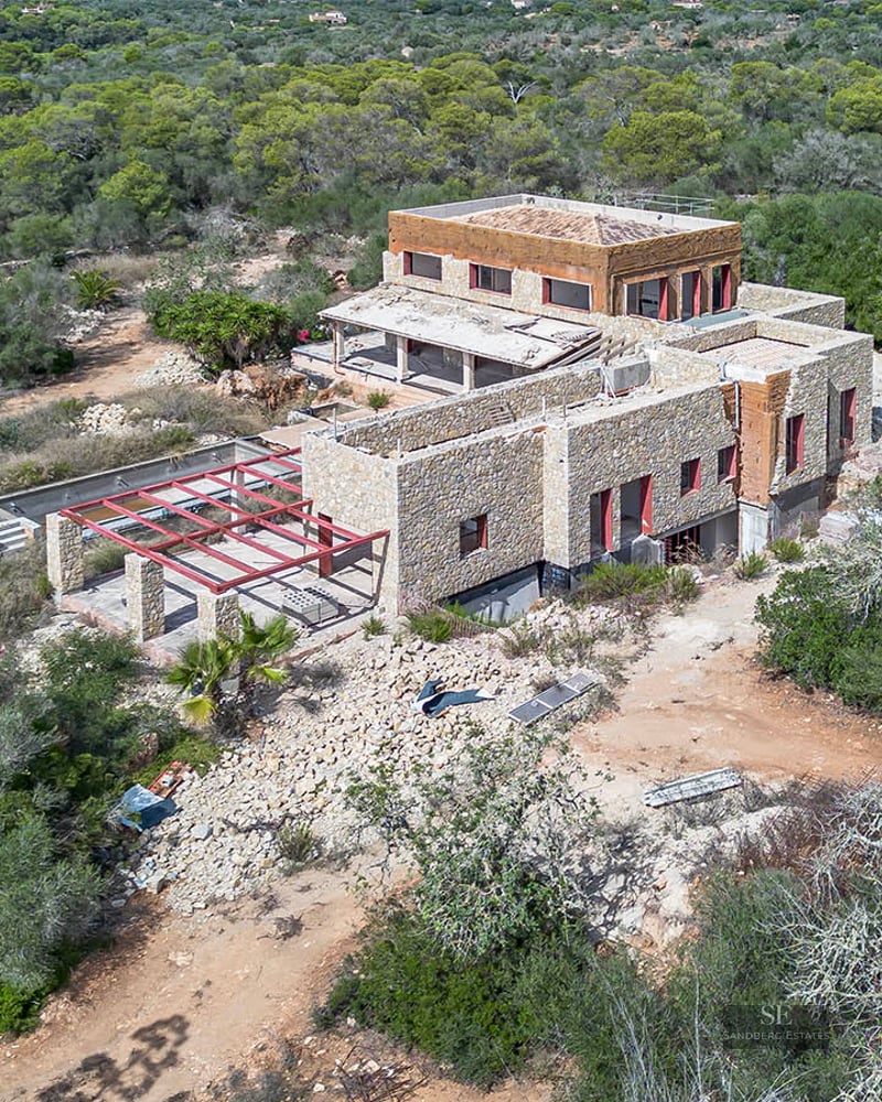 Aerial shot of a large stone villa under construction surrounded by dense Mediterranean greenery.