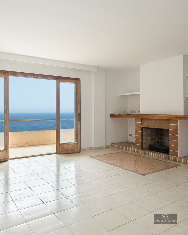 Empty white living room with a brick fireplace and wooden doors opening to a balcony with Mediterranean sea views.