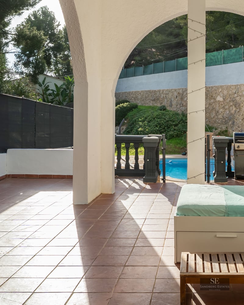 Terrasse couverte avec arches blanches, lit de repos, barbecue et vue sur une piscine ensoleillée.
