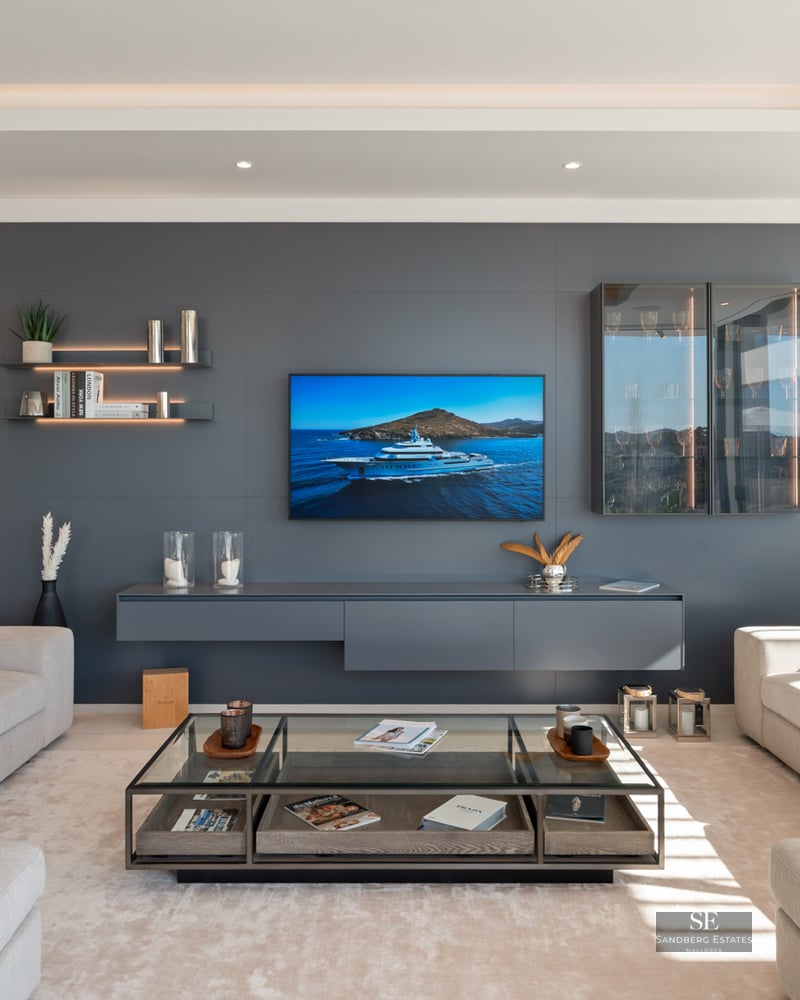 A bright, modern living room with cream sofas, a large grey media wall with TV, and a glass coffee table.