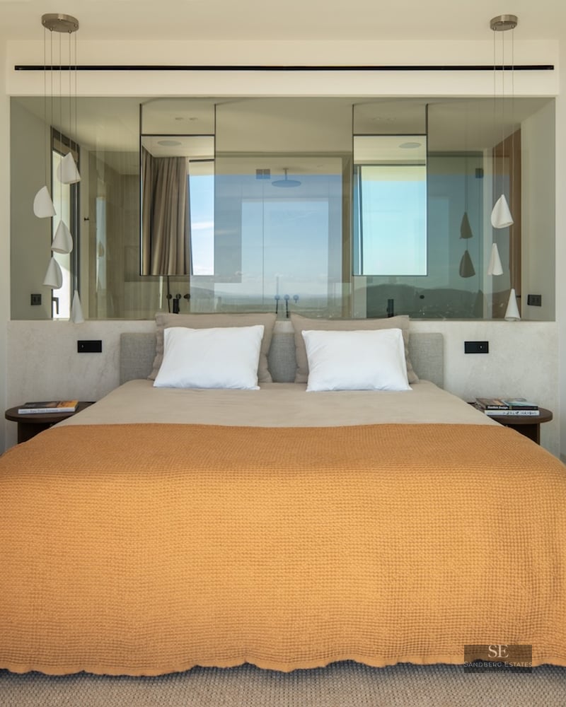 Modern bedroom featuring an orange bedspread, glass partition wall behind the bed, and sculptural white pendant lamps.