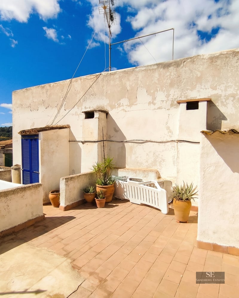 A sunny rooftop terrace with terracotta tiles, white stucco walls, potted plants, and a bright blue door.