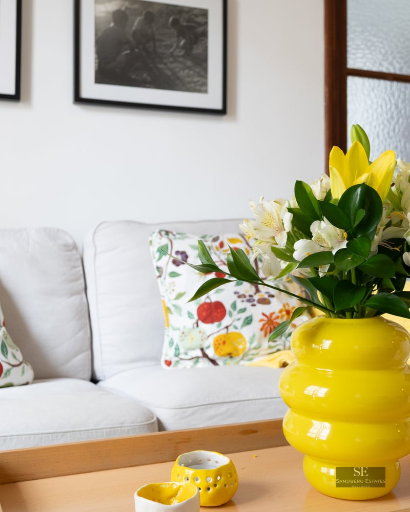 Bright living room detail with white sofa, floral pillows, and a vibrant yellow vase on a wooden coffee table.