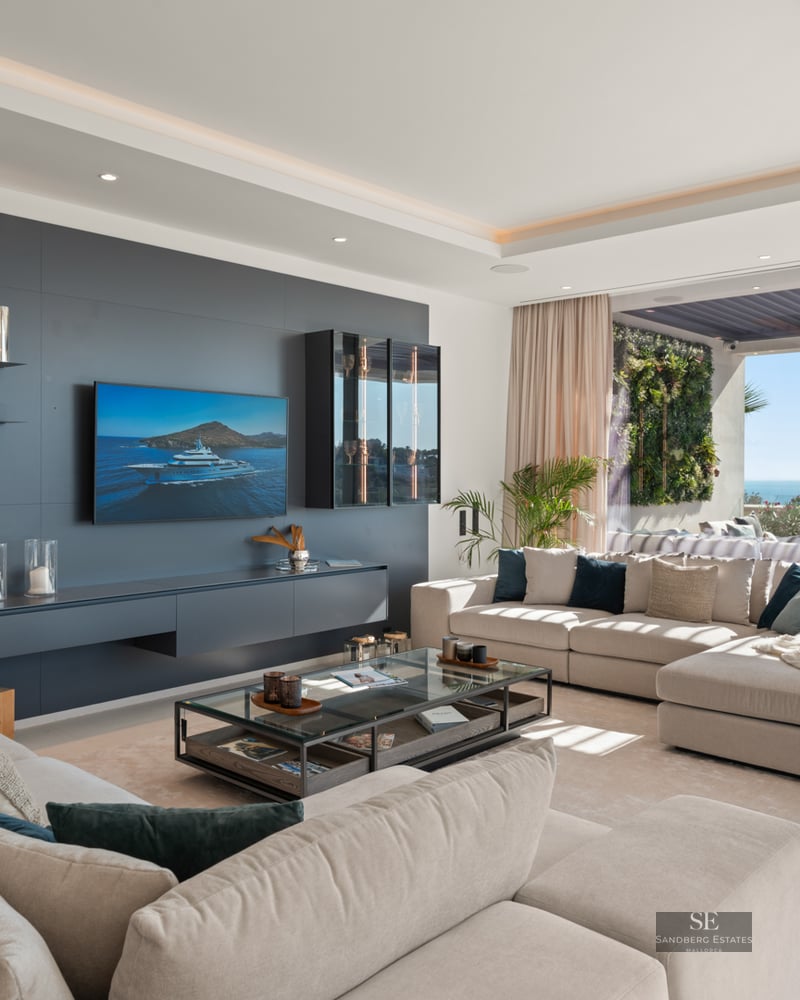 Bright living room with large beige sofas, charcoal accent wall, TV, and panoramic glass doors opening to a sea view terrace.