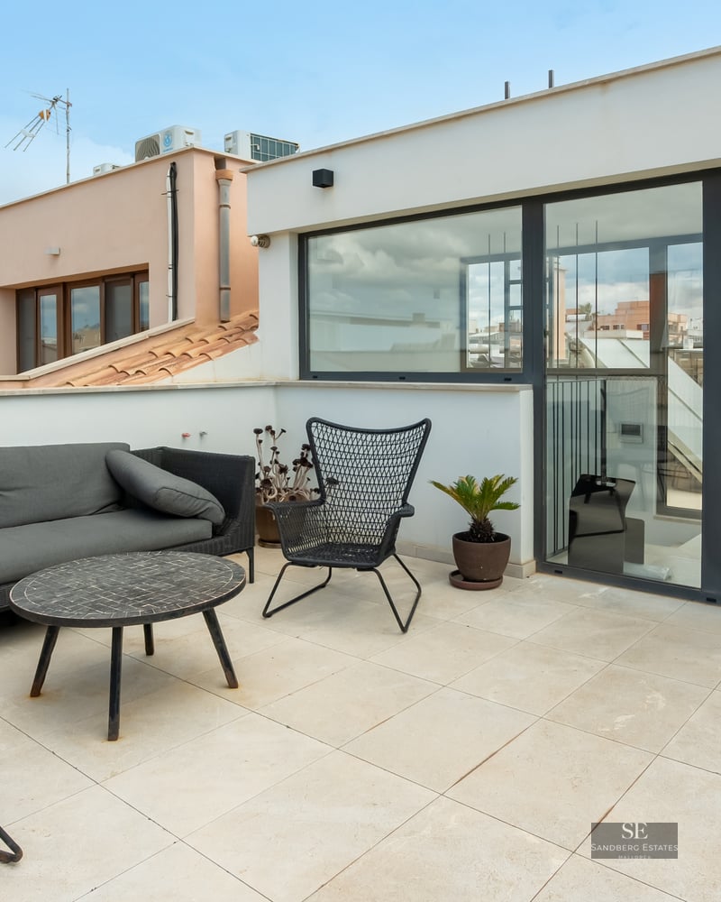 Rooftop terrace featuring grey sofa, black design chairs, and large glass doors with urban city views.