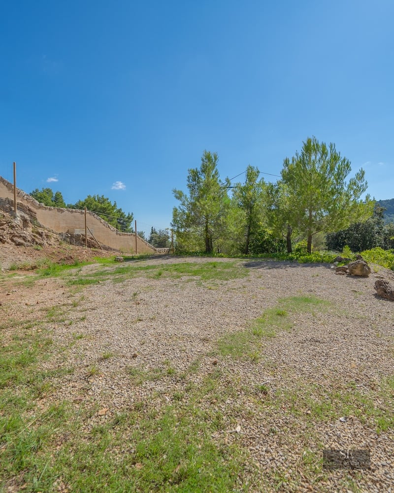 A flat gravel and grass area surrounded by pine trees and a stone wall under a clear blue sky.
