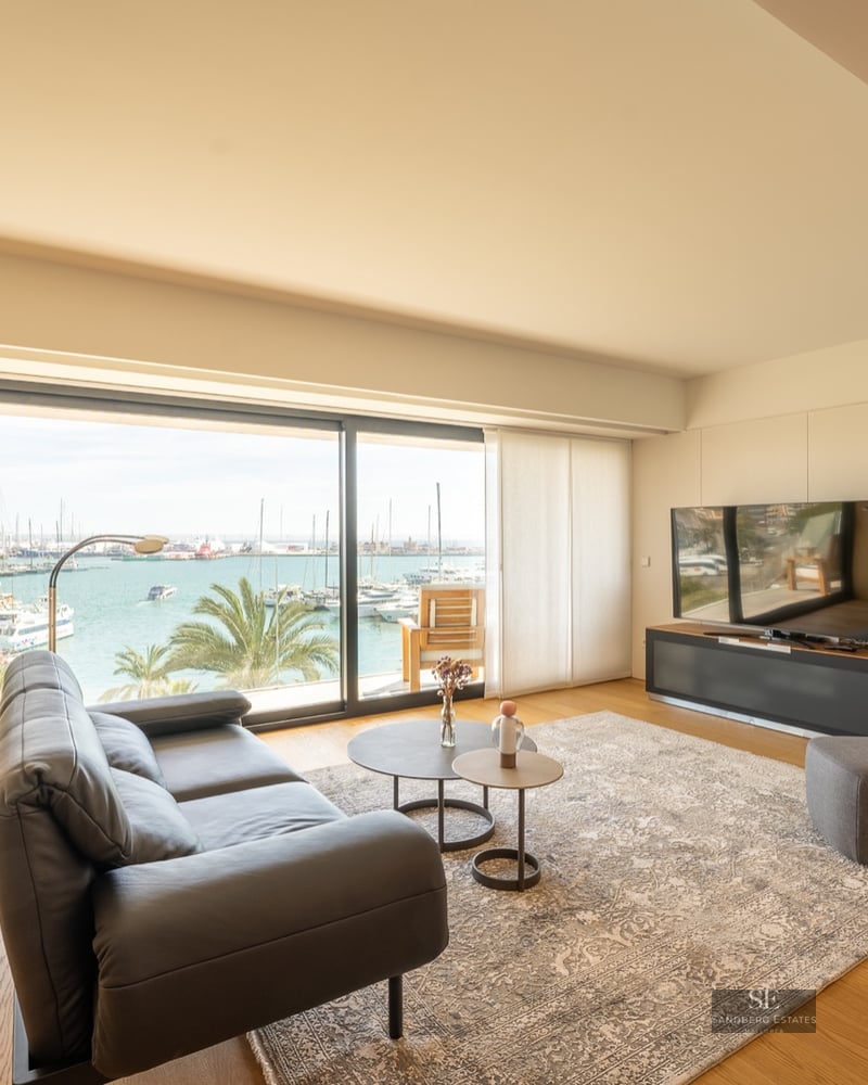 Modern living room with leather sofa and large glass windows overlooking a harbor with boats and palm trees.