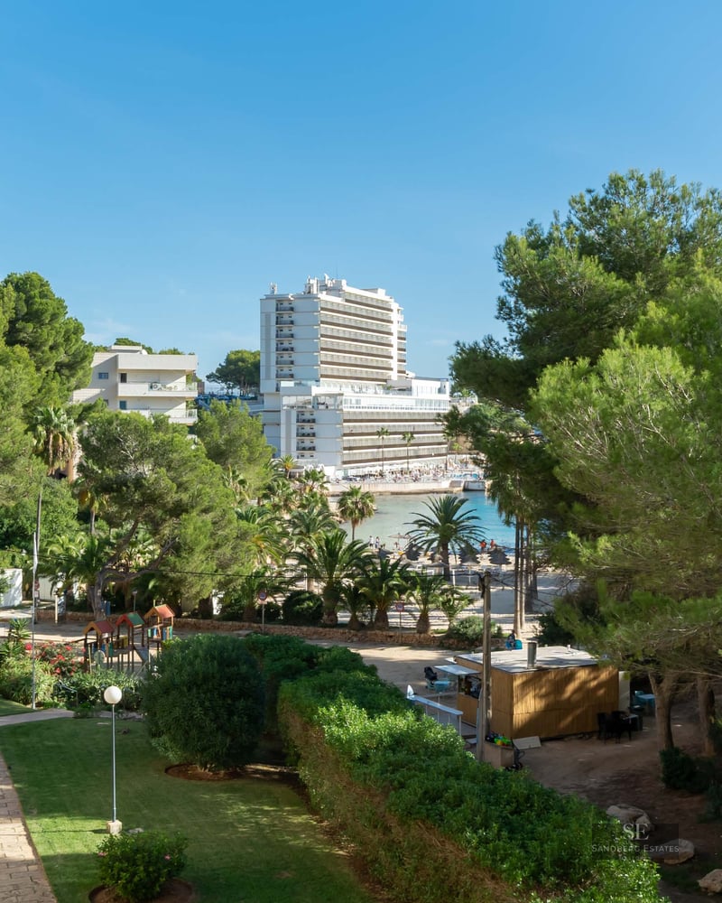 Jardin vert soigné avec des sentiers en pierre, des pins et un grand hôtel surplombant une baie sous un ciel bleu.