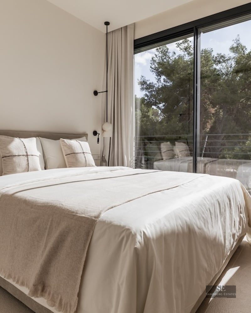 Bright bedroom with neutral-toned bed facing a large floor-to-ceiling window overlooking trees.