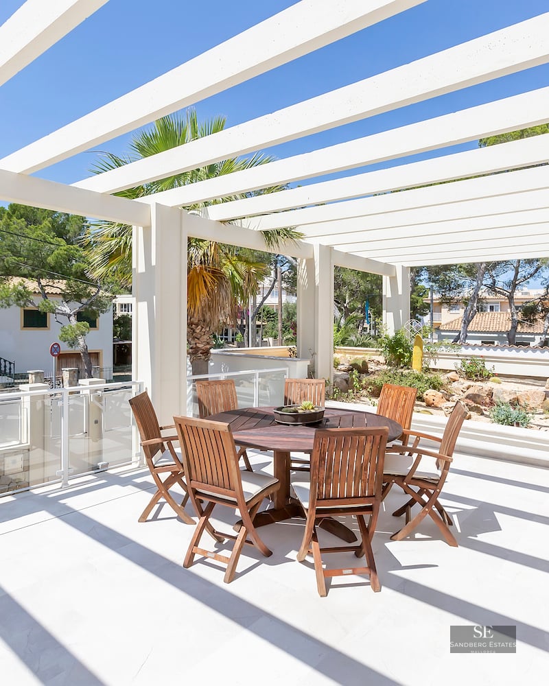 Sunny terrace featuring a white architectural pergola, round wooden dining set, and palm trees under a clear blue sky.
