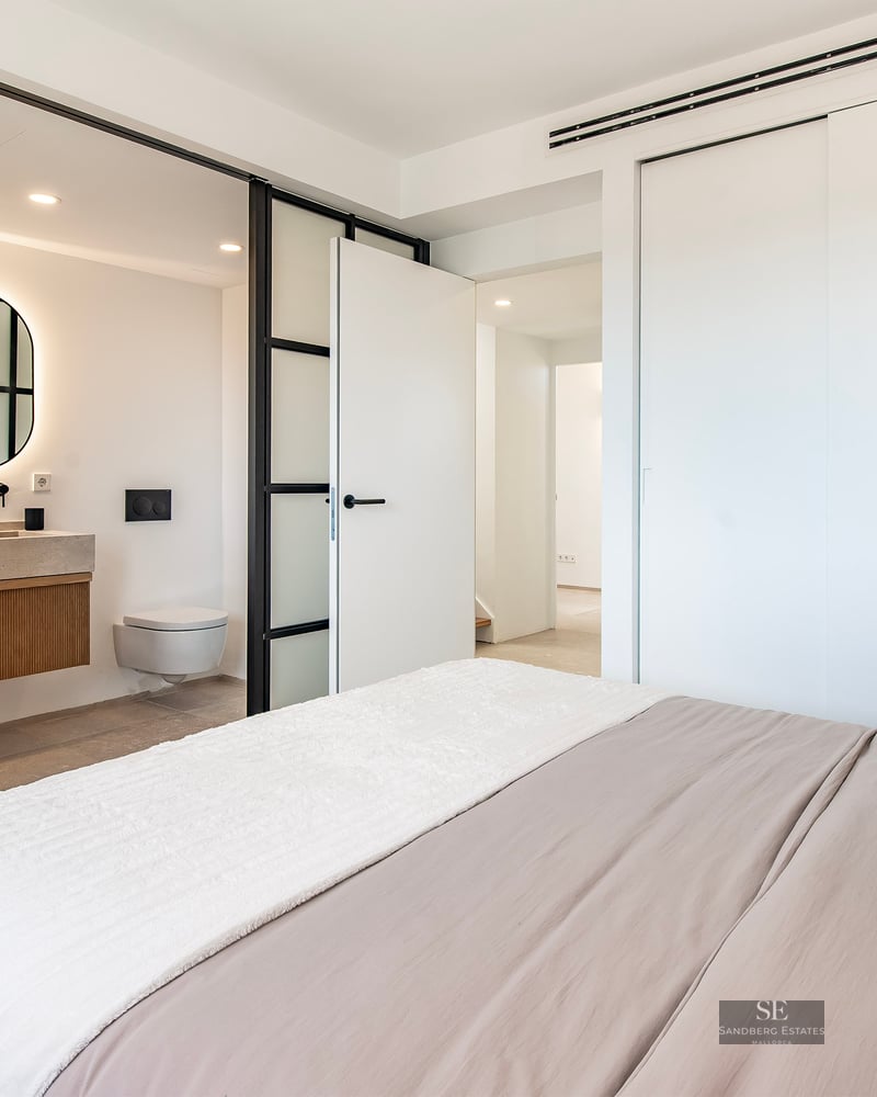 Modern bedroom featuring a bed with neutral linens and a glass-partitioned ensuite bathroom with a backlit round mirror.