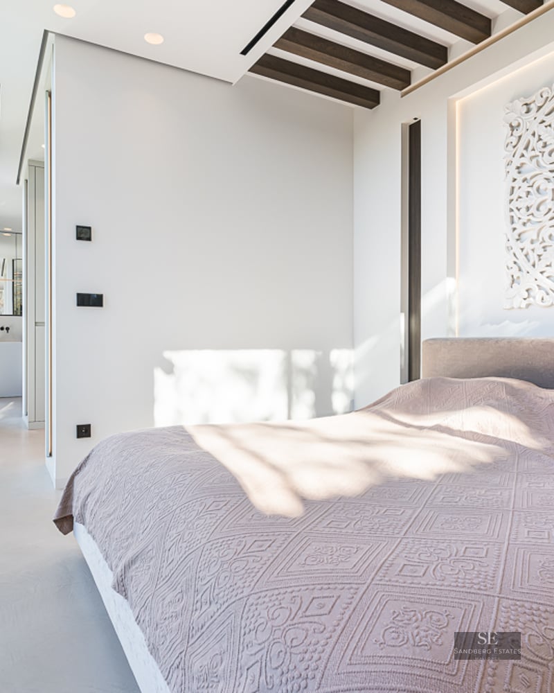 Bright modern bedroom with a large bed, floor-to-ceiling glass doors, and a decorative white carved headboard.