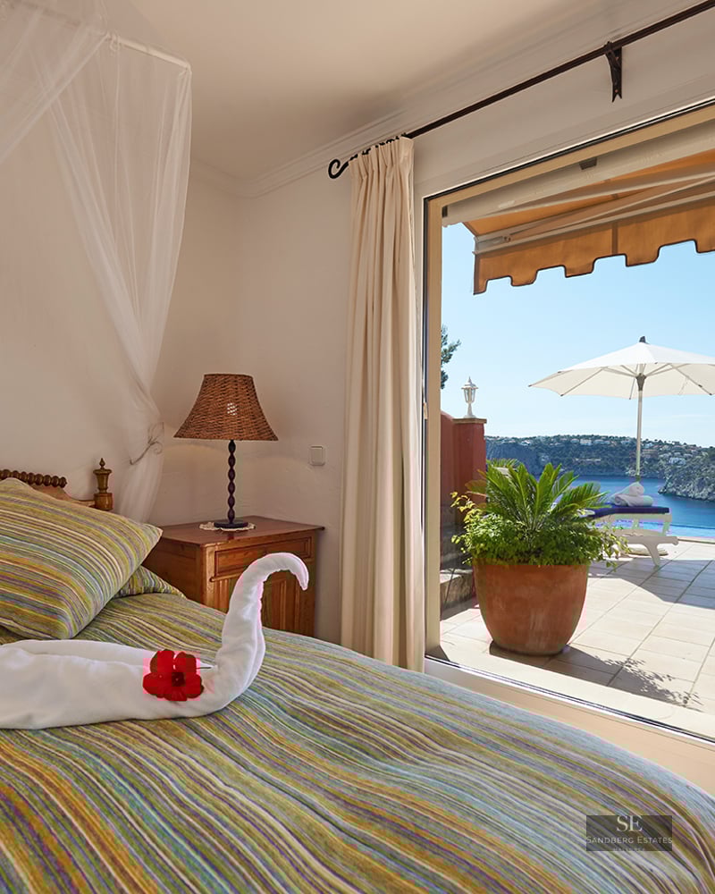 Bedroom with a striped bed and swan towel overlooking a terrace with an infinity pool and blue ocean bay.