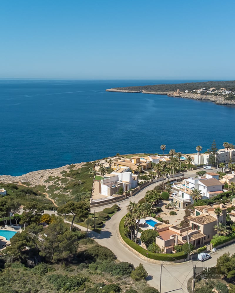 Aerial view of luxury white villas with blue pools along a rocky Mediterranean coastline under a clear blue sky.