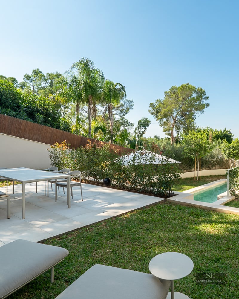 Modern white terrace featuring a dining area, outdoor grill, and lap pool surrounded by lush landscaping and palm trees.