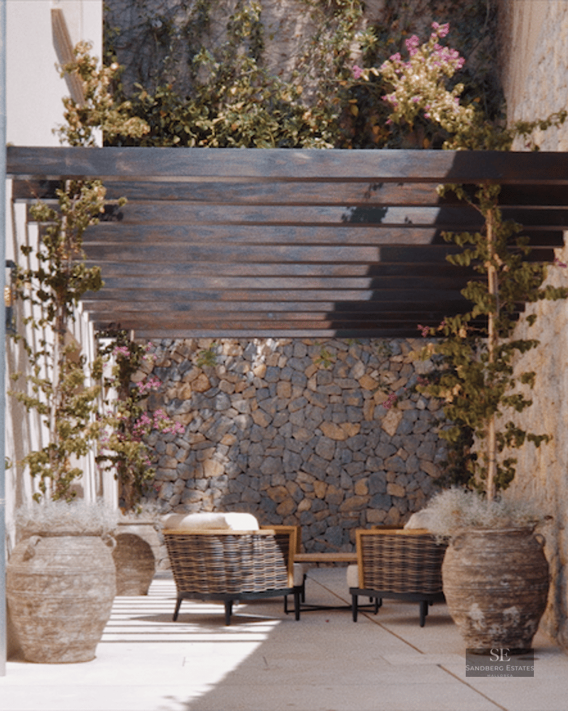 Sunlit Mediterranean terrace featuring rustic stone walls, a dark wood pergola, and comfortable outdoor seating.