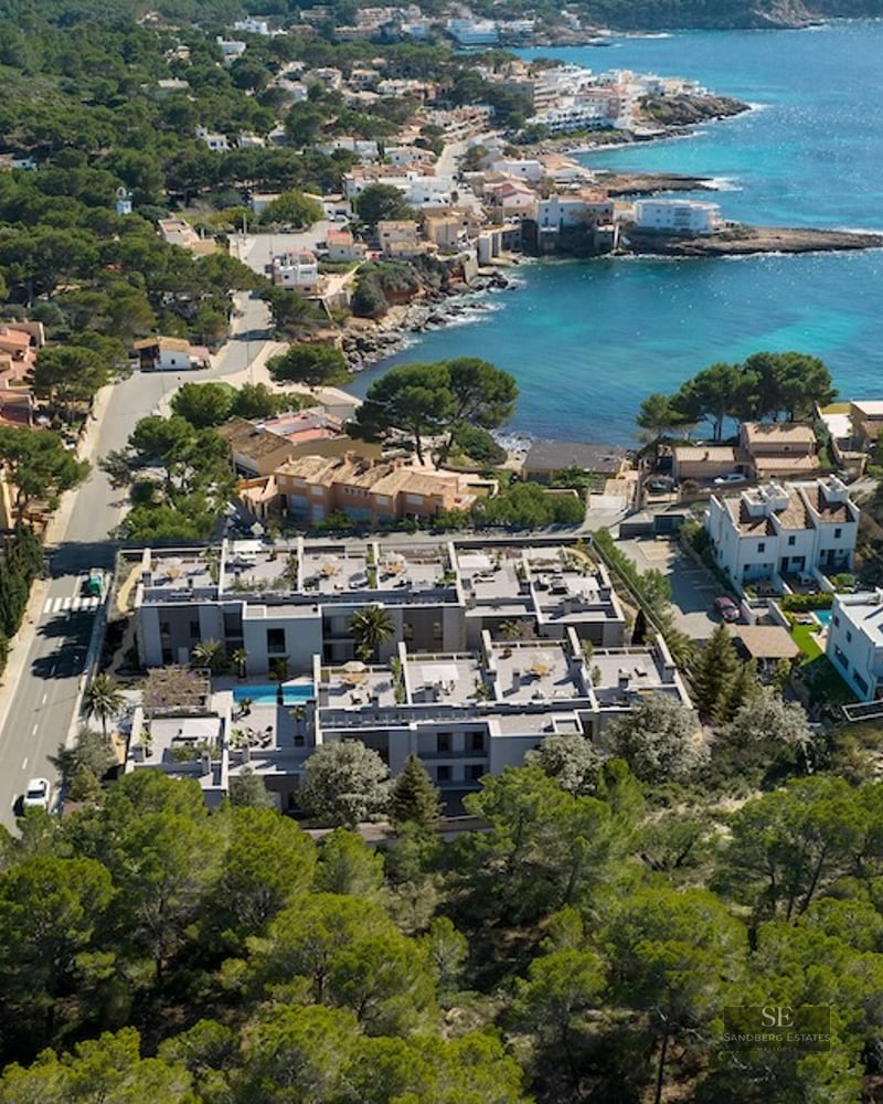 Drone view of a modern gray apartment complex nestled between pine forests and a turquoise Mediterranean bay.