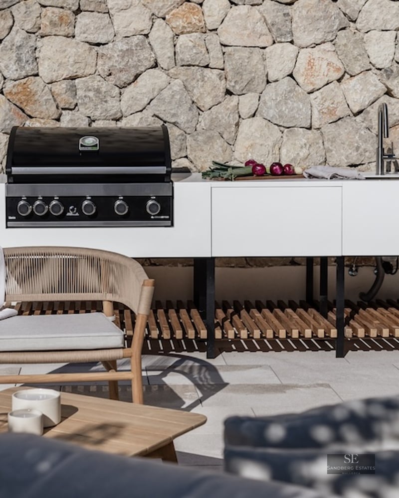 Modern white outdoor kitchen with gas grill and sink against a stone wall with designer patio furniture.