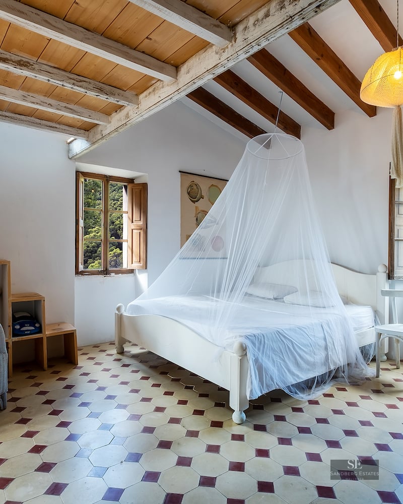 Bedroom featuring white walls, exposed wooden ceiling beams, patterned tile floor, and a bed with a mosquito net.