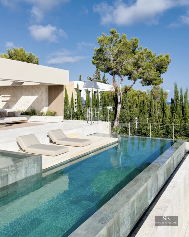 Large pool with sun loungers, surrounded by gardens and a modern luxury villa with large windows and balconies. Minimalist design.